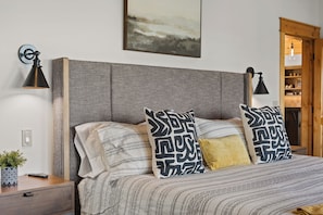 Ein Bett mit gemusterten Betttextilien und dekorativen Kissen, eine Nachttischlampe, ein kleiner Topfpflanze und ein an der Wand befestigtes Kunstwerk.