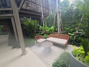 A modern outdoor seating area with wicker furniture and a round table, surrounded by lush greenery and a building in the background. - Pet-Friendly Phuket Apartment | Pool & Gym (Cherngtalay)