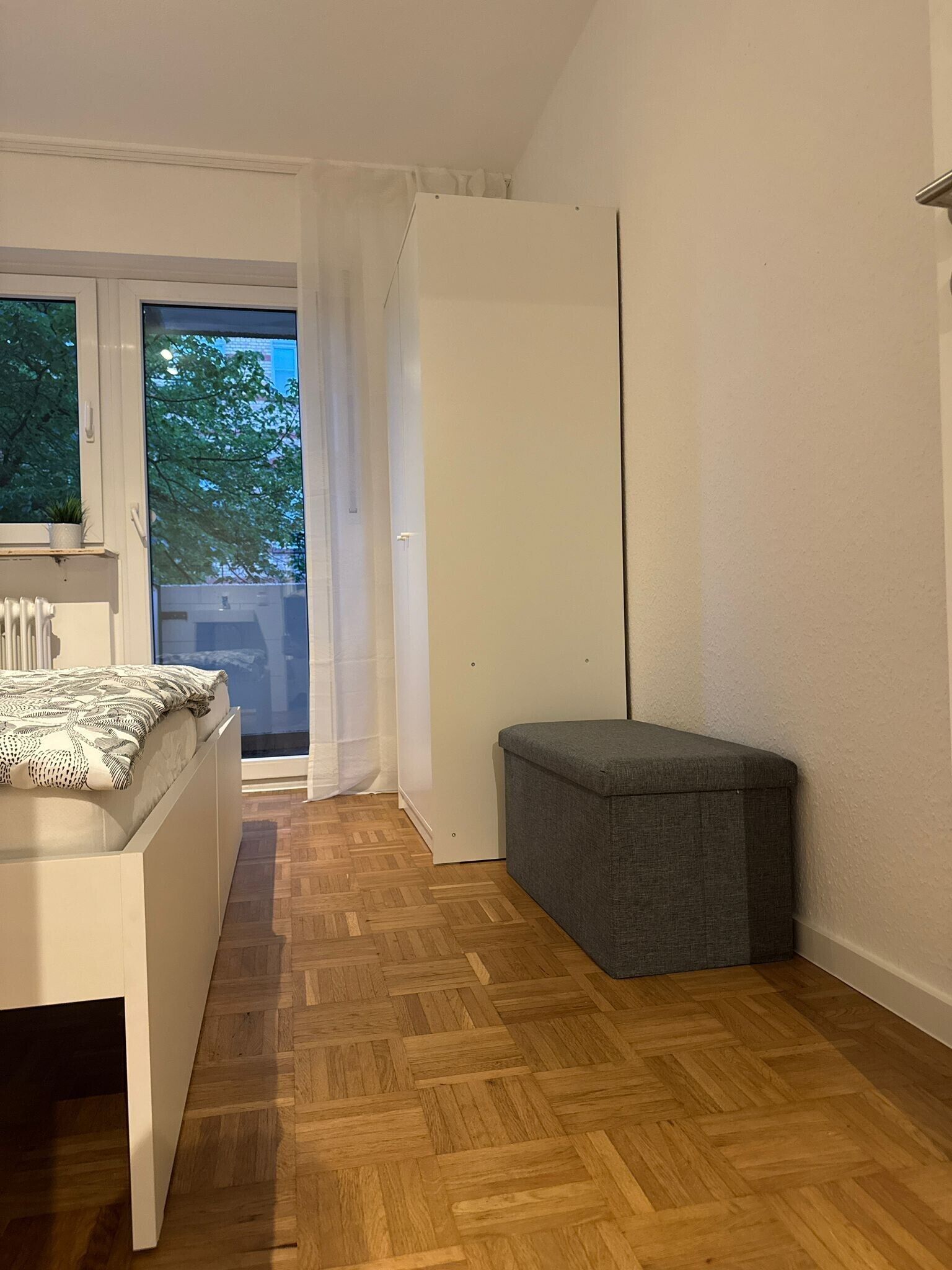 Ein Schlafzimmer mit einem Bett, einem grauen Hocker, einem Heizkörper und einem Fenster mit Blick auf Bäume.