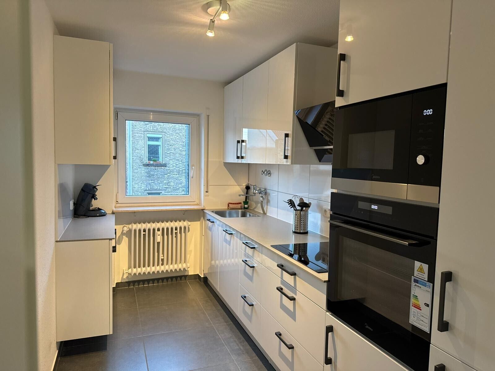 A modern kitchen with white cabinets, a built-in oven, and a window overlooking a stone building.
