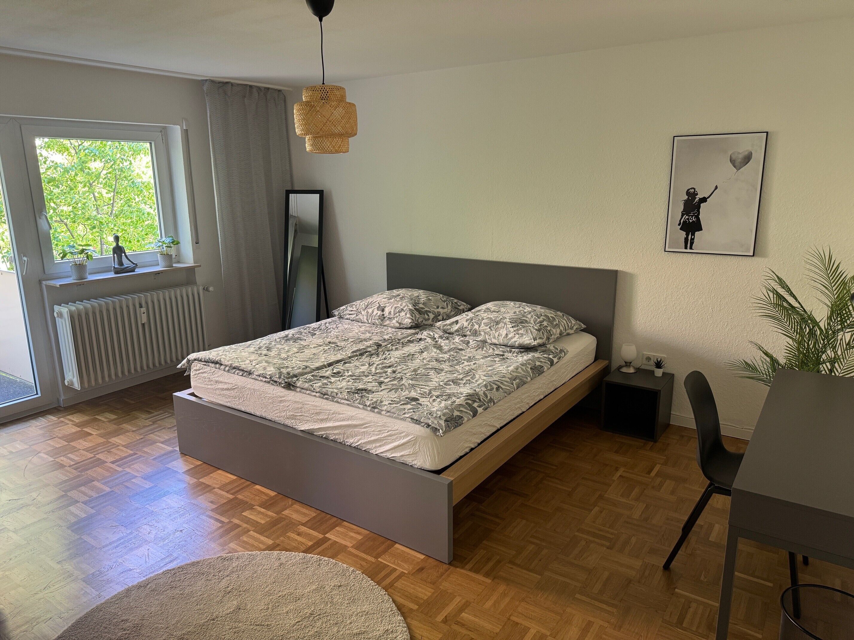 Une chambre à coucher avec un lit, un radiateur, une fenêtre avec des rideaux, un luminaire suspendu, un tableau encadré au mur et une plante.