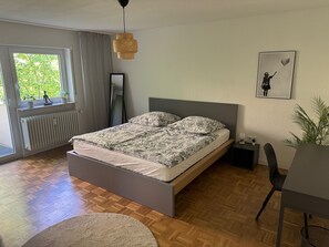 Ein Schlafzimmer mit Bett, Heizkörper, Fenster mit Vorhängen, einer Pendelleuchte, einem gerahmten Bild an der Wand und einer Pflanze.