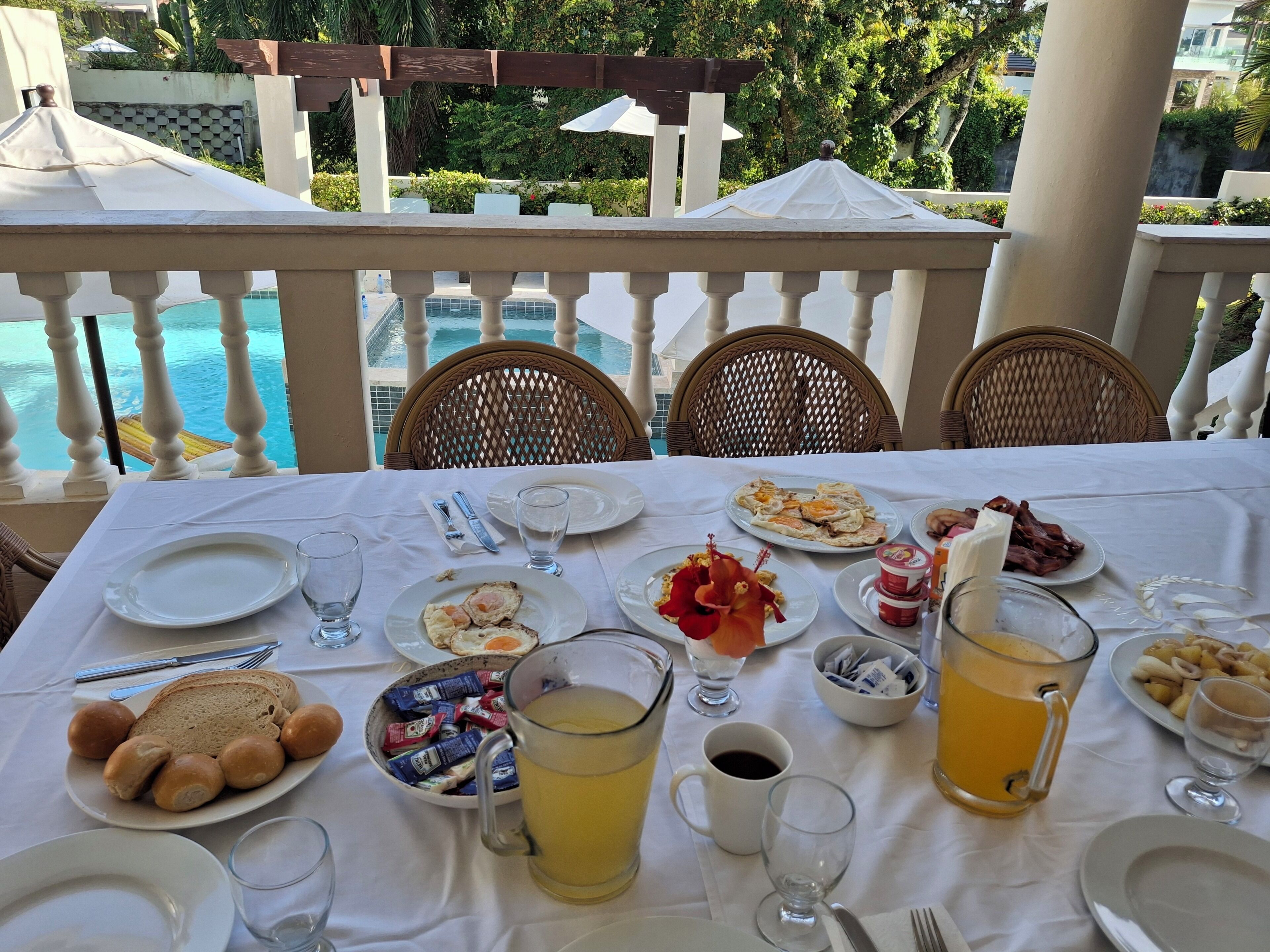 Mesa lista para el desayuno con platos, vasos y una jarra de jugo, con vista a una piscina.