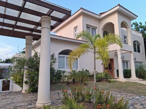 A two-story building with a covered patio area, columns, and a garden with various plants and flowers. - Your Dream Family or Group Getaway: 5 Bedroom Luxury Villa Awaits in Cofresi! (Cofresi)