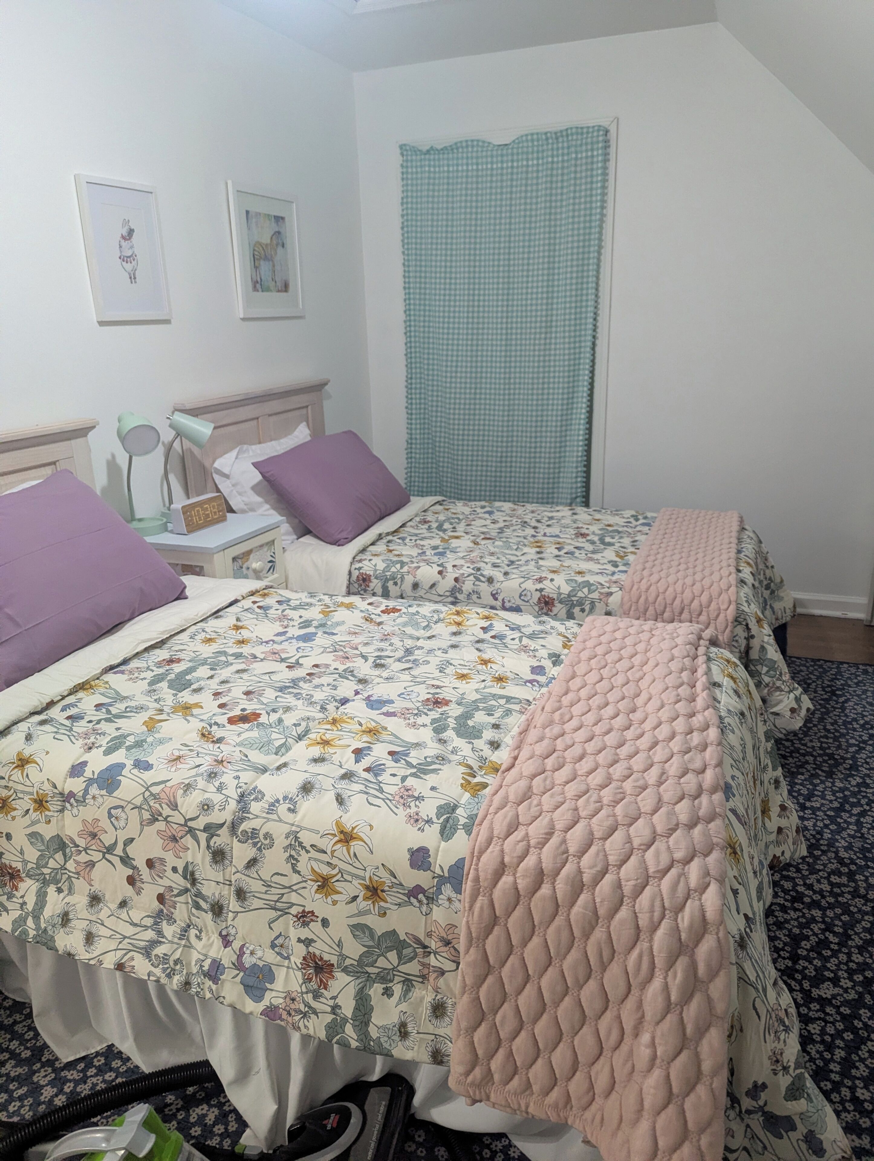 A bedroom with two beds, a nightstand, a lamp, a clock, and a window with a checkered curtain.