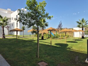 Eine gepflegte Rasenfläche mit reetgedeckten Sonnenschirmen, Palmen und einem weißen Gebäude im Hintergrund.