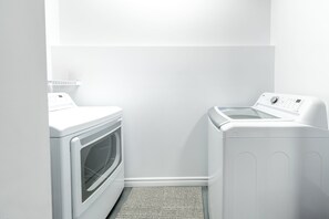 A laundry room with a front-loading washer and dryer, a small shelf above the dryer, and a textured mat at the entrance. - Spa & Nature | Chalet Le WeSki (Saint-Philémon)