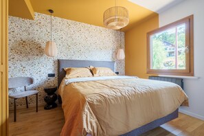 Ein Schlafzimmer mit einem Bett, einem Sessel, einem Nachttisch, einem Fenster und einer Wand mit blumigem Tapetenmuster.