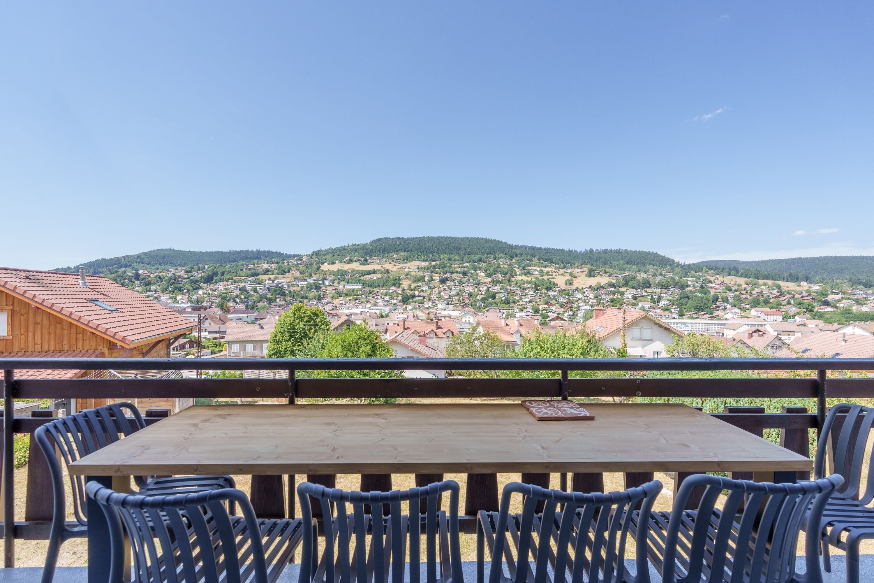 Mesa de madeira com cadeiras numa varanda com vista para uma cidade com casas e colinas.