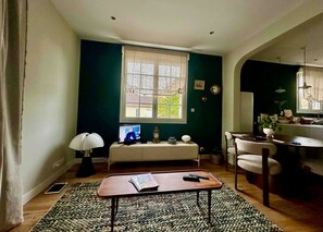 A living room with a television, a small table, a sofa, and a window with curtains. - Villa de la Hève - Rénovée, 700 m de la plage (Ouistreham)