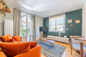 A modern living room with a sofa, a TV, a dining area with a table set, and a large window with curtains. - Villa de la Hève - Rénovée, 700 m de la plage (Ouistreham)