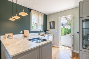 A modern kitchen with a marble countertop, pendant lights, a window with curtains, and a view of the outside. - Villa de la Hève - Rénovée, 700 m de la plage (Ouistreham)