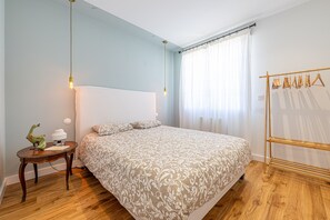 Un dormitorio con una cama grande, una mesita de noche con lámpara, suelo de madera y una ventana con cortinas.