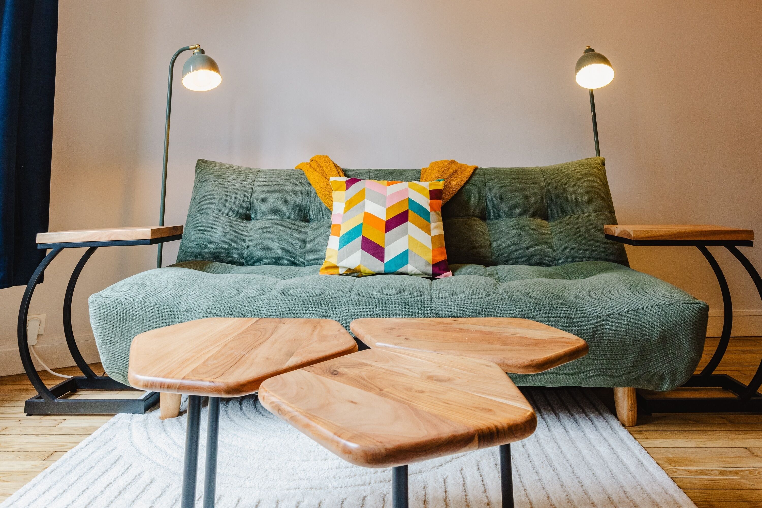A green sofa with a colorful chevron pillow, two wooden side tables, and two floor lamps.