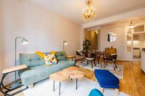Ein modernes Wohnzimmer mit einer Couch, zwei Beistelltischen, einem Couchtisch und einer blauen Bank. Ein Essbereich mit Tisch und Stühlen ist vorhanden, und im Hintergrund ist eine Küche zu sehen.