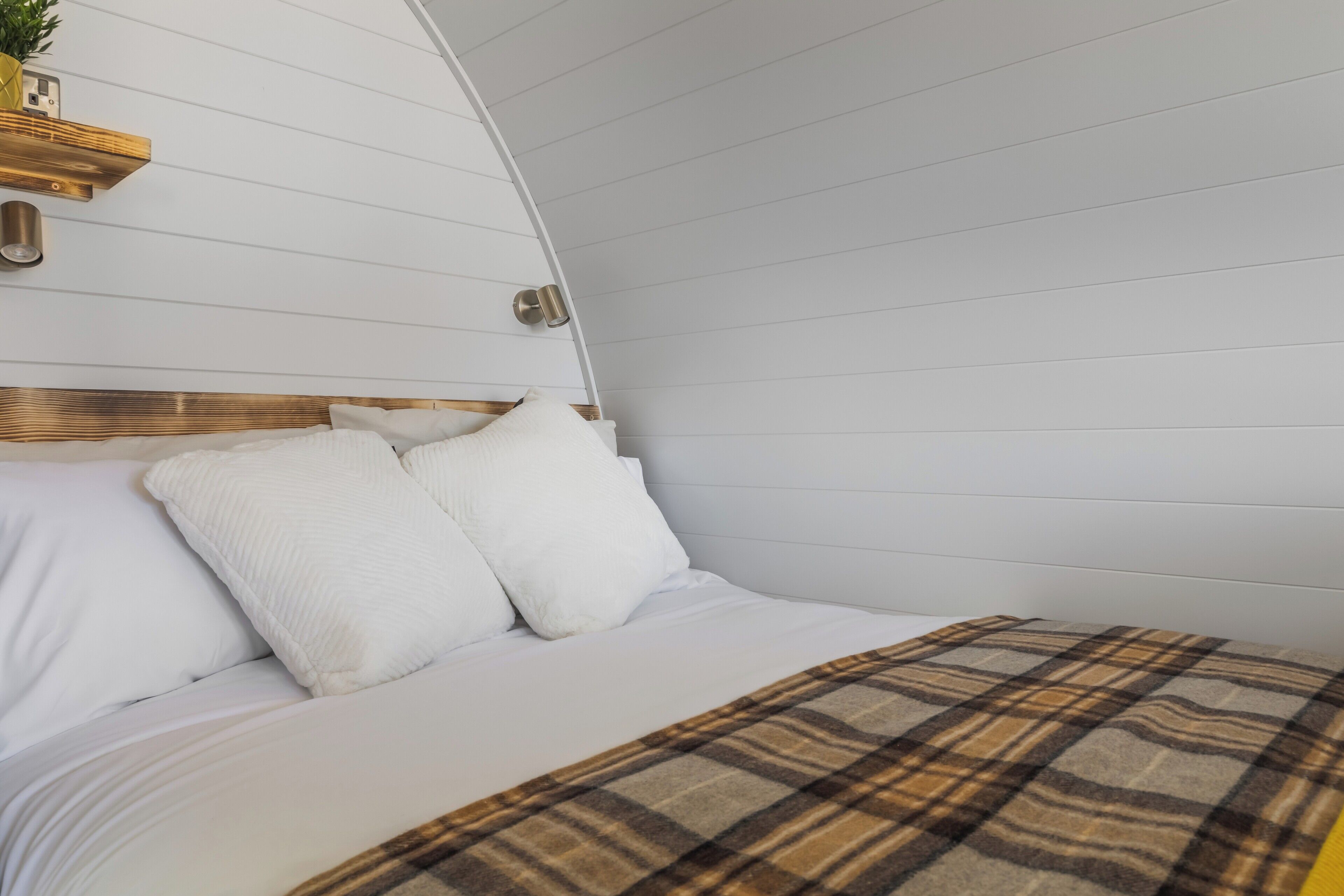 Uma cama com roupa de cama branca e manta xadrez, cabeceira de madeira e luminária fixada na parede.