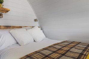 Una cama con ropa de cama blanca y una manta escocesa, cabecera de madera y una lámpara fijada a la pared.