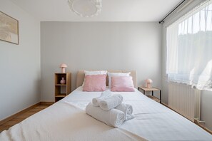 Een net opgemaakt bed met witte lakens en opgerolde handdoeken, een houten nachtkastje met een roze lamp, en een bijzettafeltje met een vergelijkbare lamp.