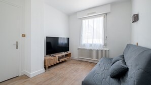 Ein modernes Wohnzimmer mit einem Flachbildfernseher auf einem Holzständer, einem grauen Sofa und einem Fenster mit leicht transparenten Vorhängen.
