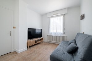 Ein modernes Wohnzimmer mit einem Flachbildfernseher auf einem Holzständer, einem grauen Sofa und einem Fenster mit leicht transparenten Vorhängen.