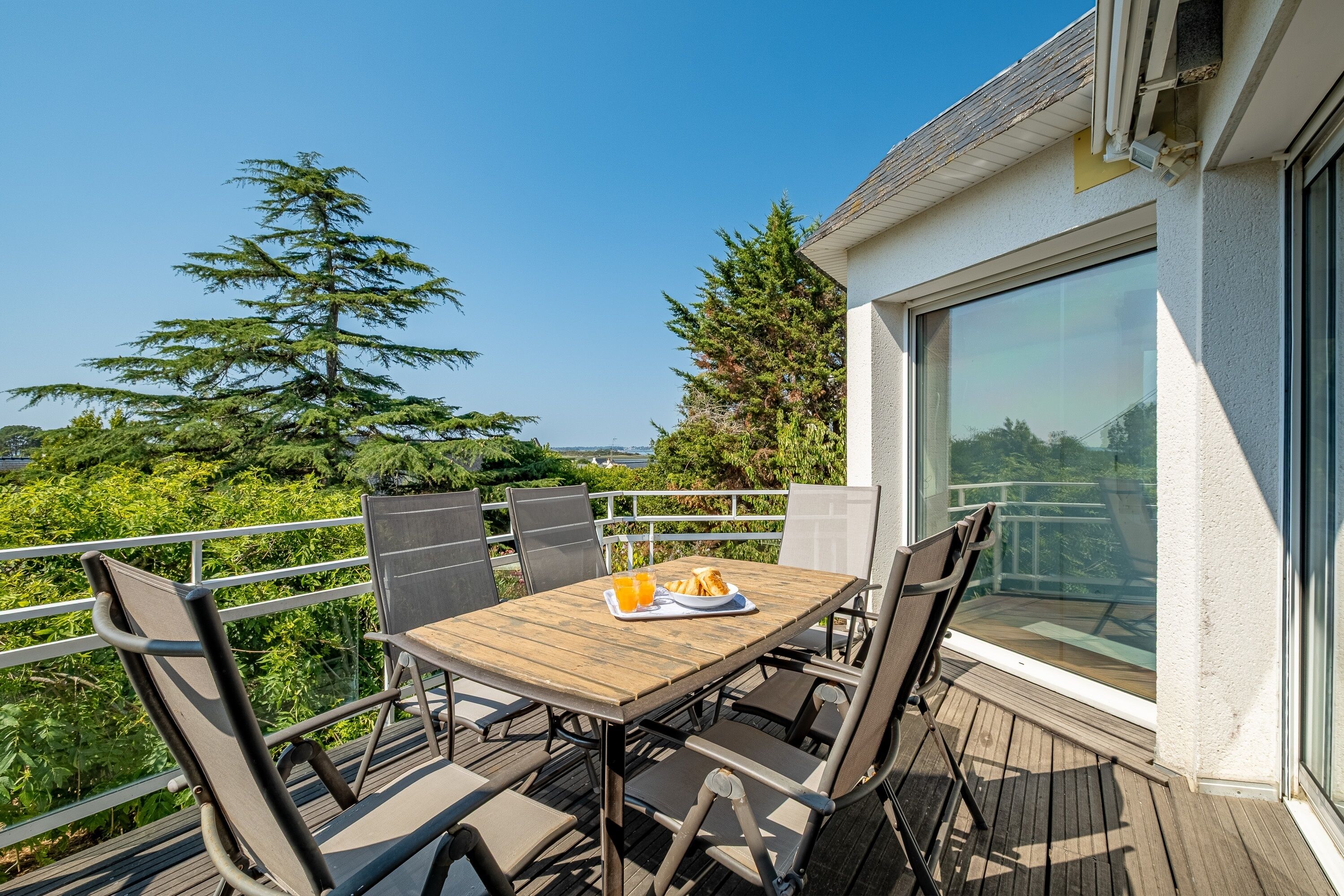 Une terrasse en bois avec une table et des chaises, offrant une vue panoramique sur un paysage verdoyant avec des arbres et un ciel dégagé.