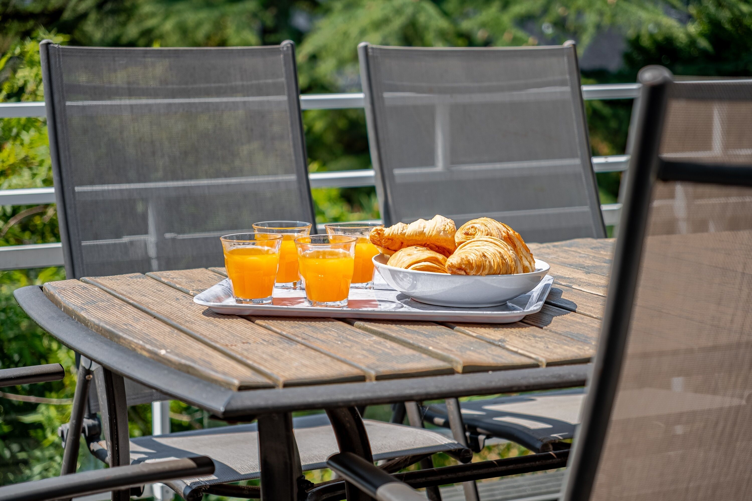 A table with breakfast items including croissants and orange juice, set outdoors with chairs and a view of greenery.