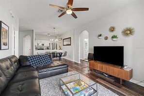 A modern living room with a leather sofa, a glass coffee table, a flat-screen TV, and a ceiling fan. - Penthouse w/ Lake View | Free Resort Access | Disney 9 min (Four Corners)