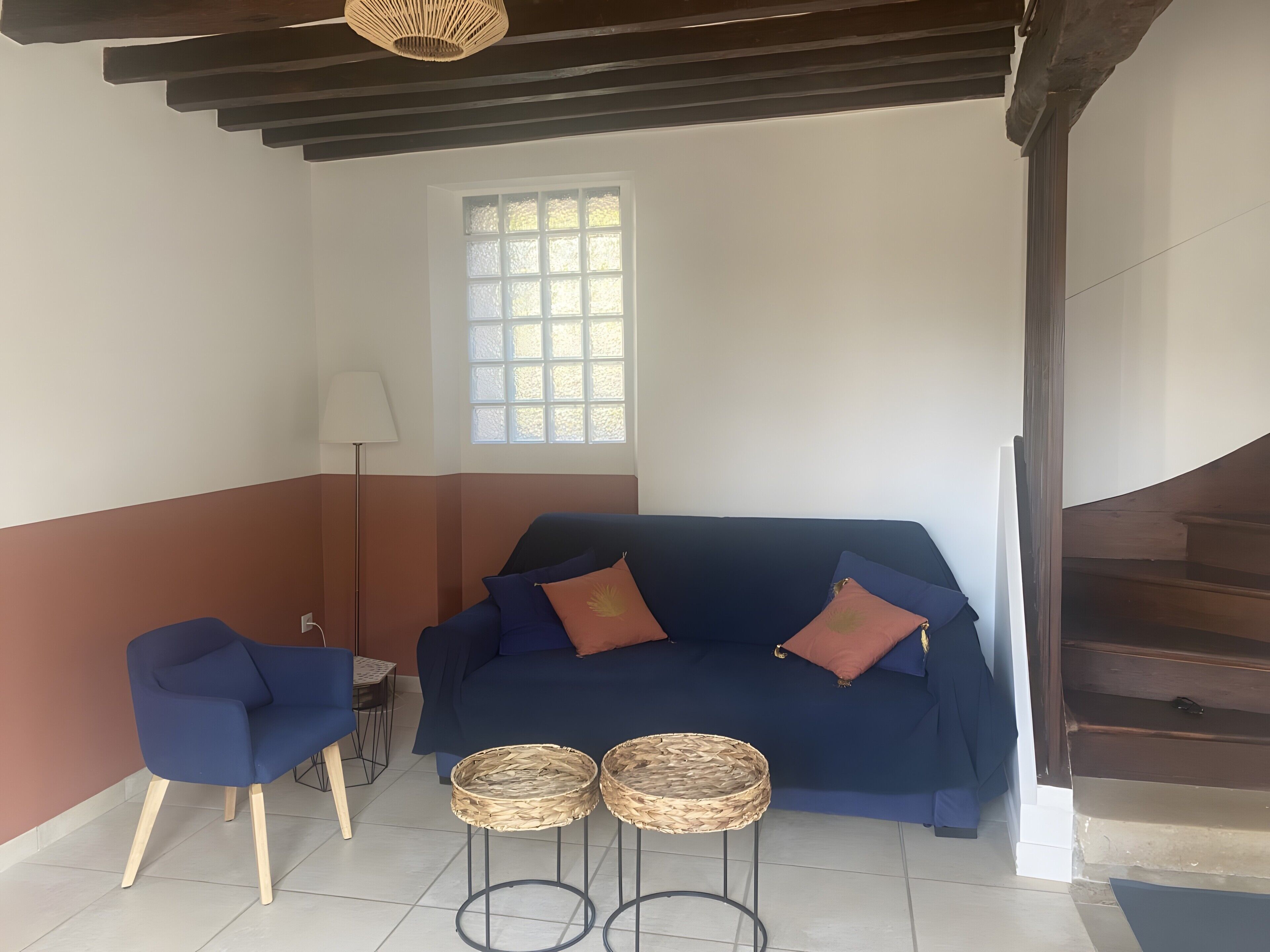 Une pièce avec un canapé bleu, deux petites tables d’appoint en osier, une lampe sur pied et une fenêtre donnant sur un beau paysage.