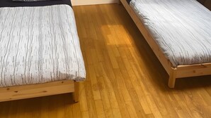 A room with two wooden beds, a small table with a lamp, and a plain wall.