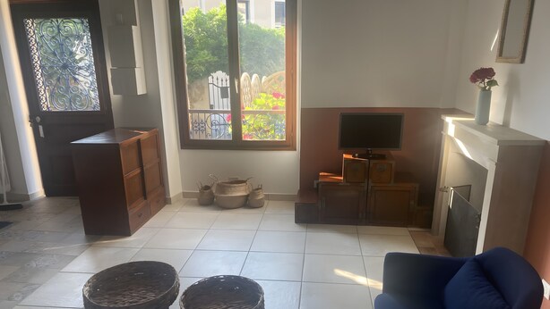 A living room with a TV, a blue armchair, a wooden cabinet, and a window with a view of greenery.