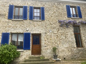 Un edificio de piedra con varias ventanas, algunas con contraventanas, una puerta de madera y una planta en maceta.