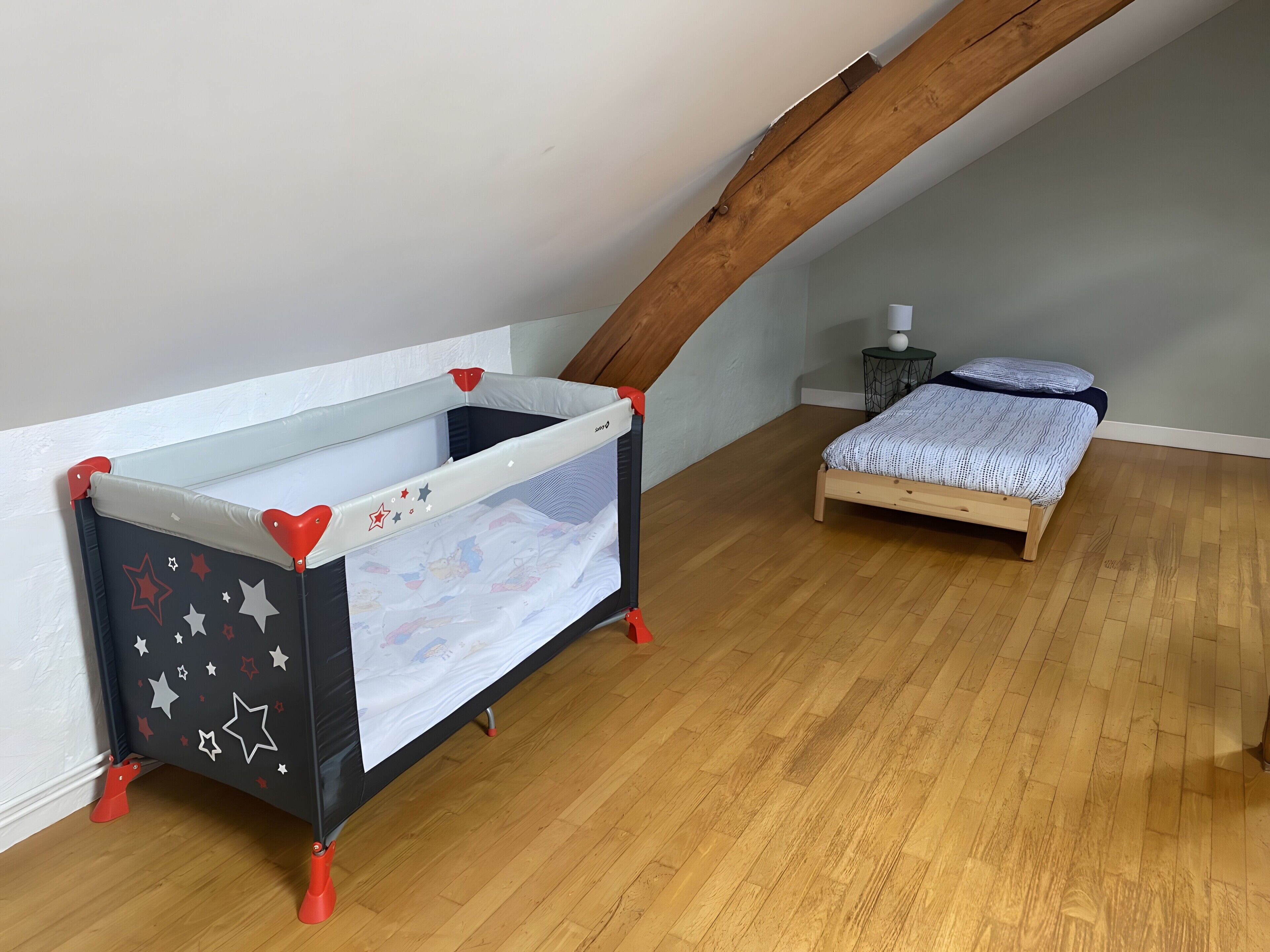 A room with a sloped ceiling, a crib with star patterns, a small bed, and a nightstand with a lamp.