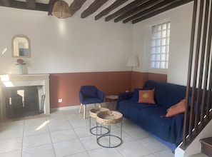Un salon avec un canapé bleu, une petite table, un fauteuil, une cheminée et une fenêtre avec des stores.