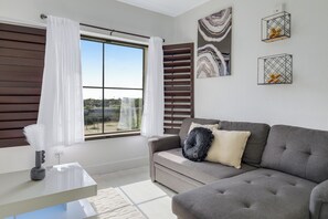 A modern living room with a grey sofa, a glass coffee table, and a view of the outdoors through a large window with white curtains.
