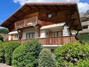 Eine zweigeschossige Holzhütte mit Balkon, Steinaußenwand und üppiger Bepflanzung.