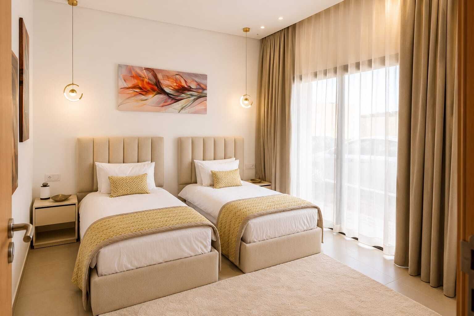 Et hotelværelse med to senge, et stort vindue med gardiner, et billede på væggen og et natbord med en lampe.