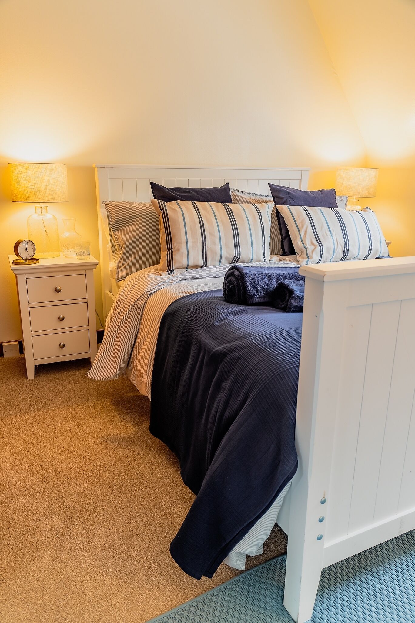 Een opgemaakt bed met streepjesbeddengoed, een nachtkastje met een lamp, en een nachtkastje met een klok.