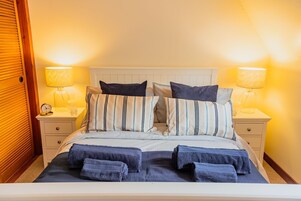 Una cama bien hecha con ropa de cama a rayas y varios almohadones, flanqueada por mesitas de noche con lámparas y un reloj.