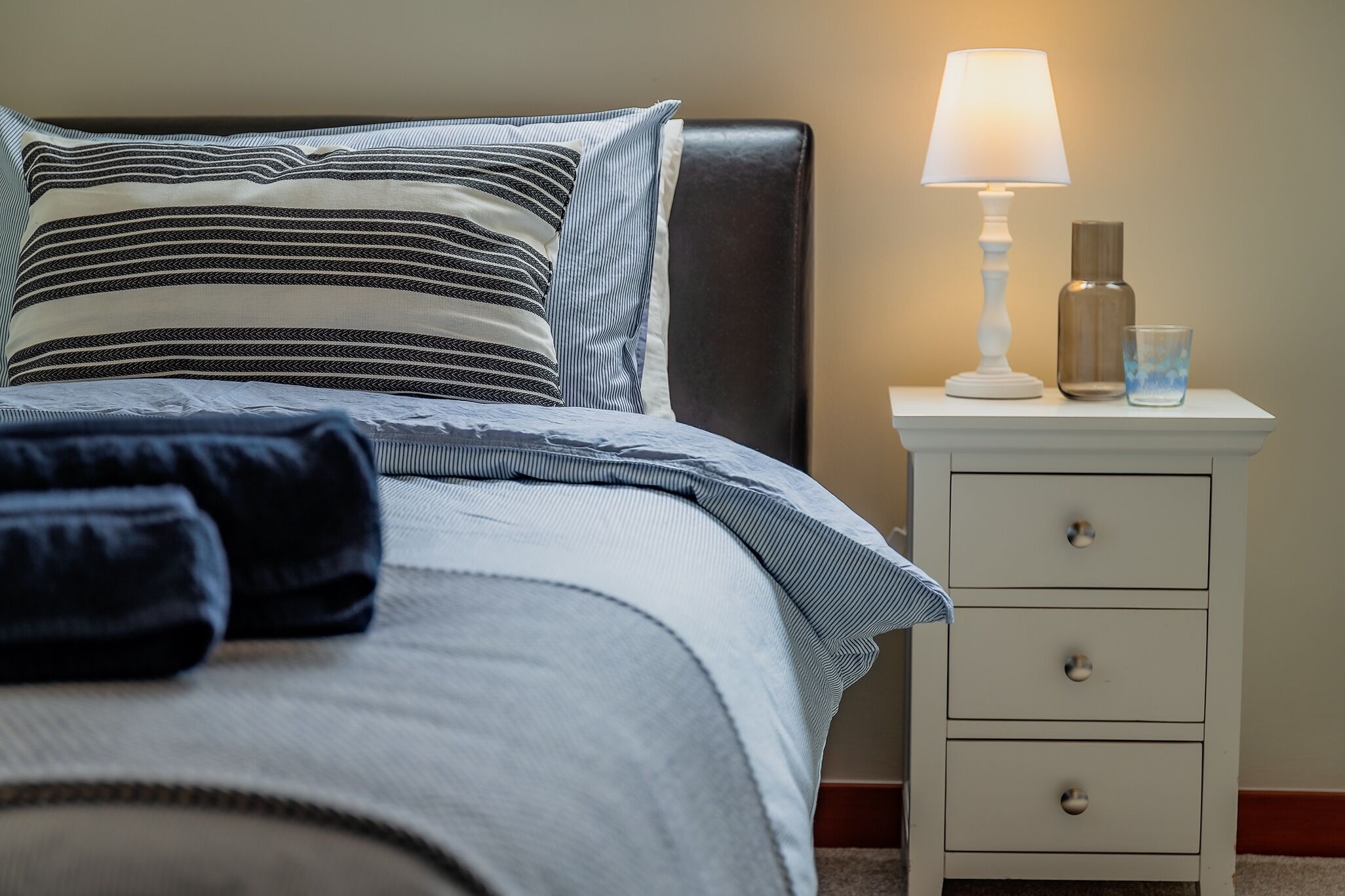 A neatly made bed with striped bedding and a pillow, a bedside lamp, a glass of water, and a white nightstand with drawers.