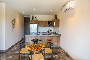 A modern kitchen with wooden cabinets, a central island, and a glass-top dining table with two chairs. - Luxury 3BR Tulum Penthouse with Private Pool (Tulum)