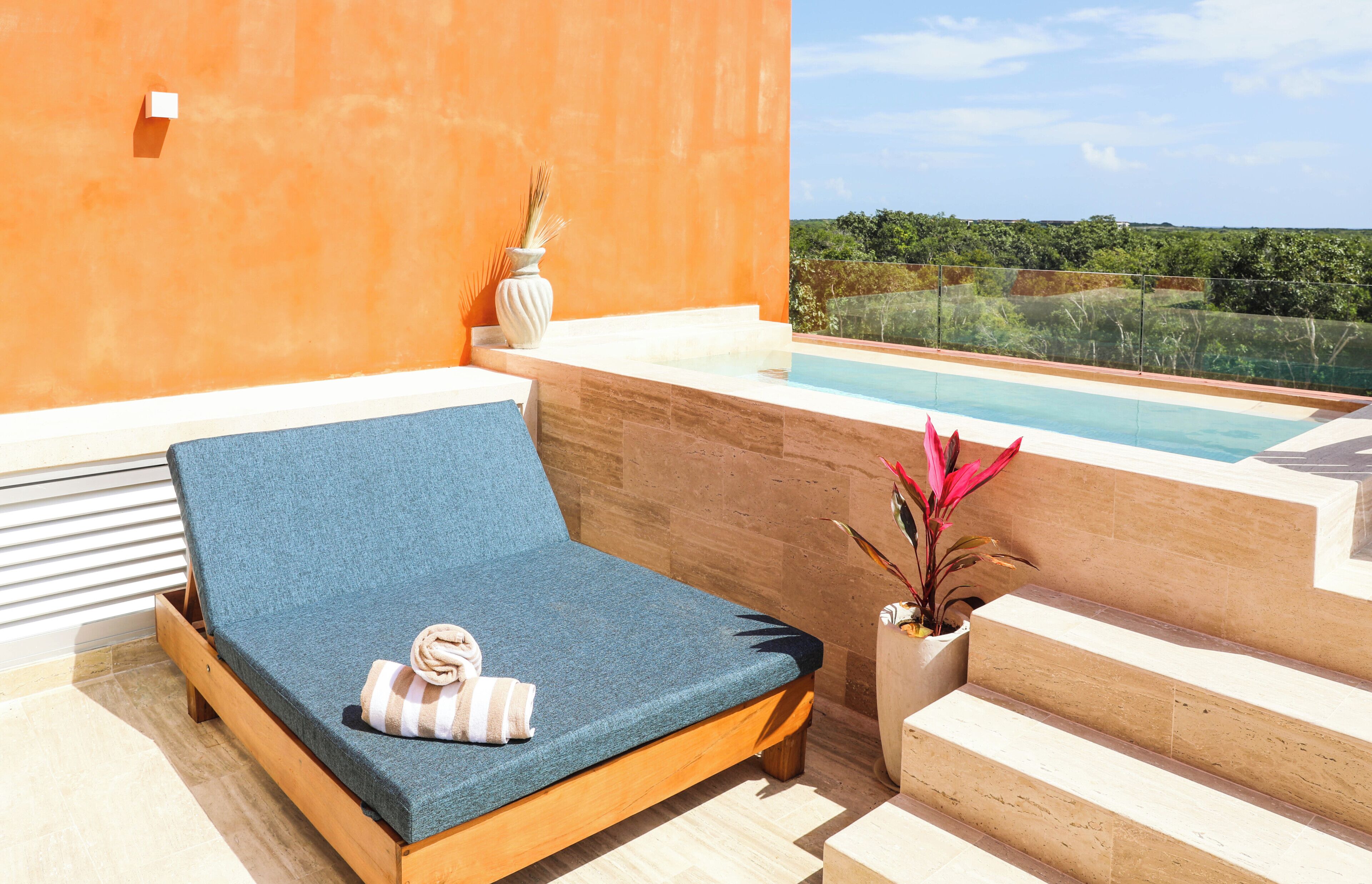 A sun lounger with a towel, a potted plant, and a view of a pool and greenery.