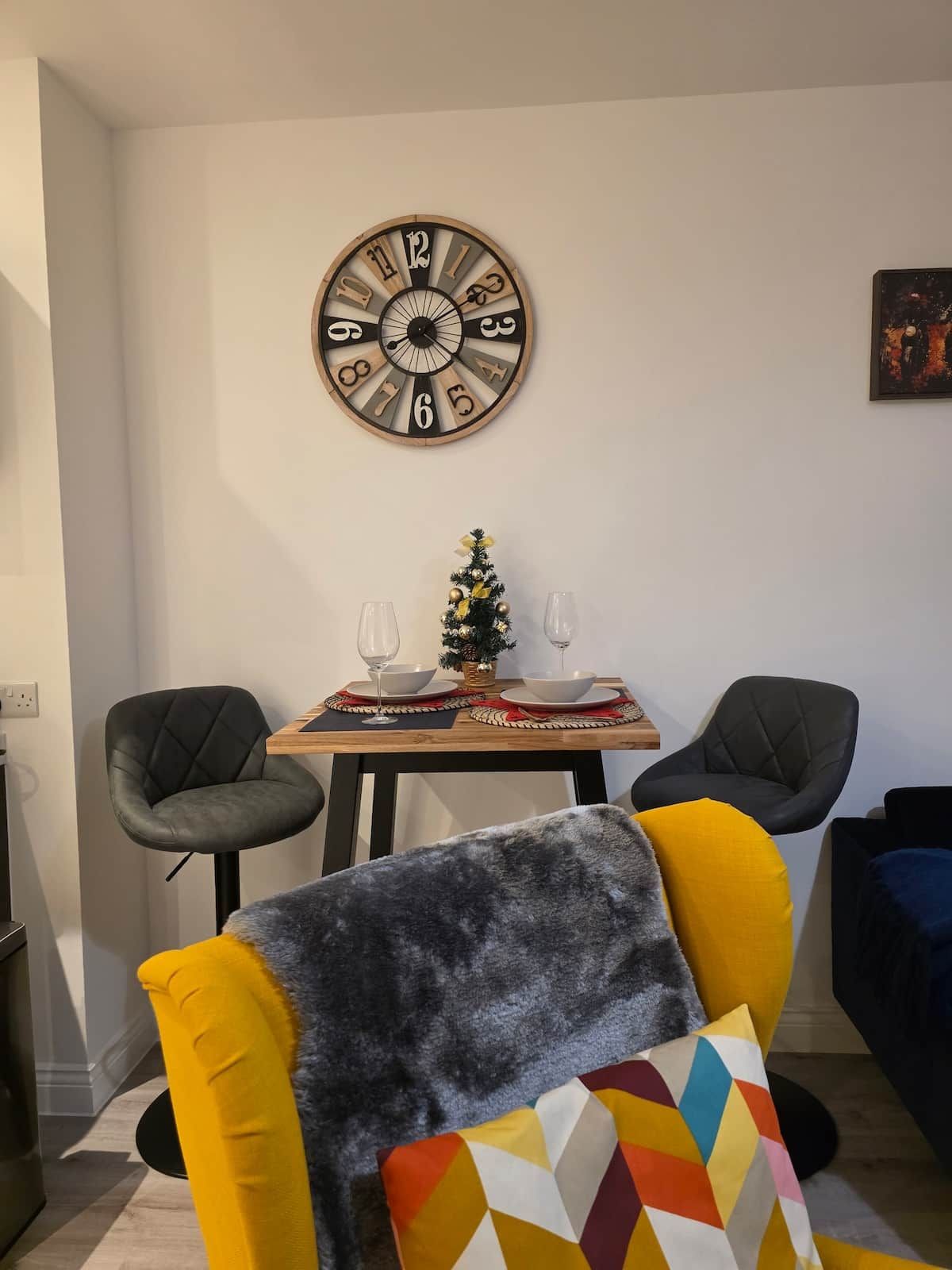 A dining area with a wooden table set for two, two chairs, a small Christmas tree, and a wall clock.