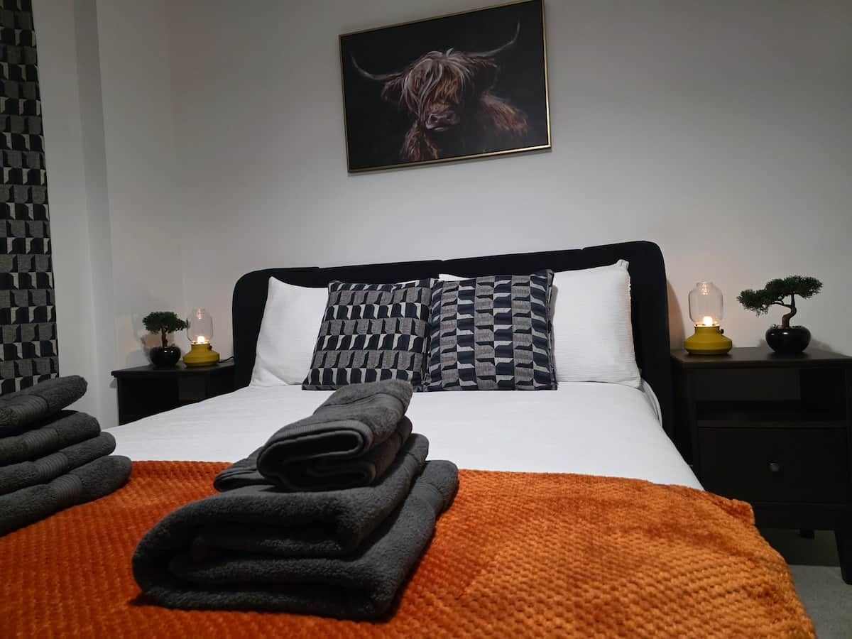 Een opgemaakt bed met een kussentje met ruitpatroon, een gevouwen handdoek en een bedlampje op een nachtkastje.