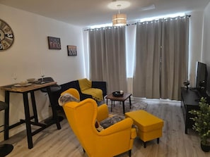Ein Wohnzimmer mit Esstisch, gelben Armlehnstühlen, einer Couch, einem Fernseher und einer Wanduhr.