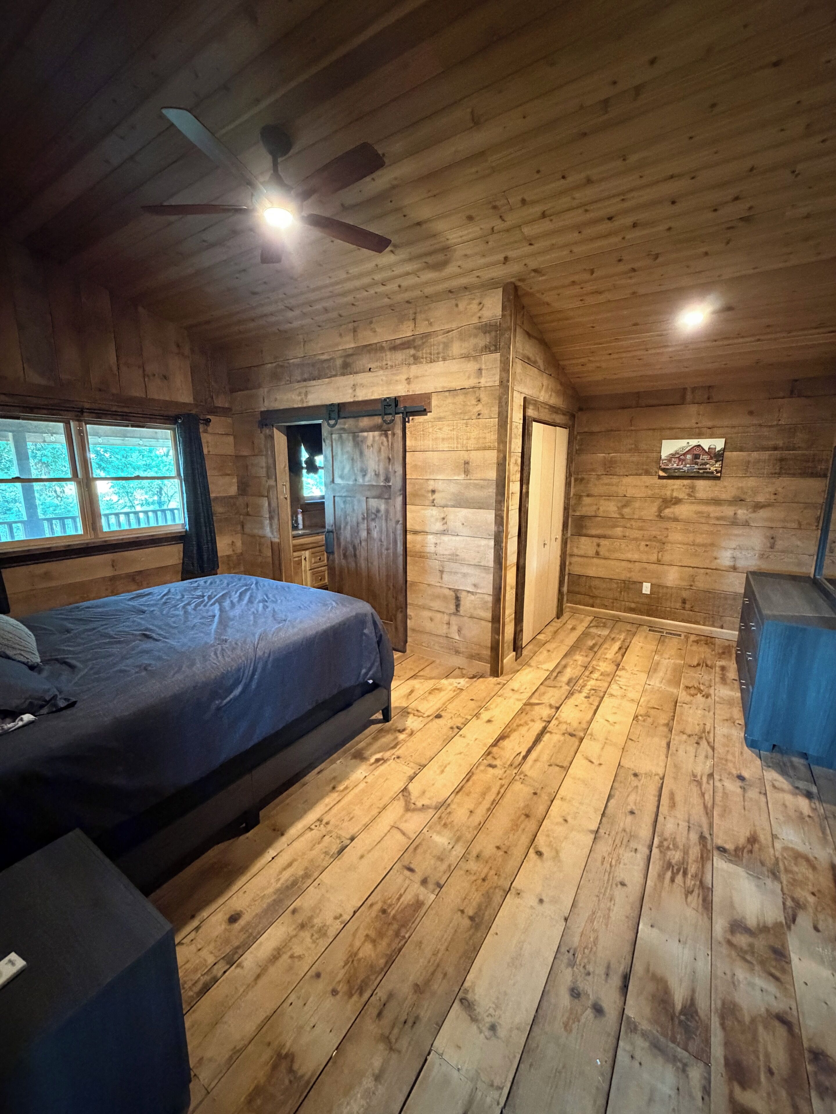 A wooden cabin bedroom with a bed, a ceiling fan, a window, and a door.