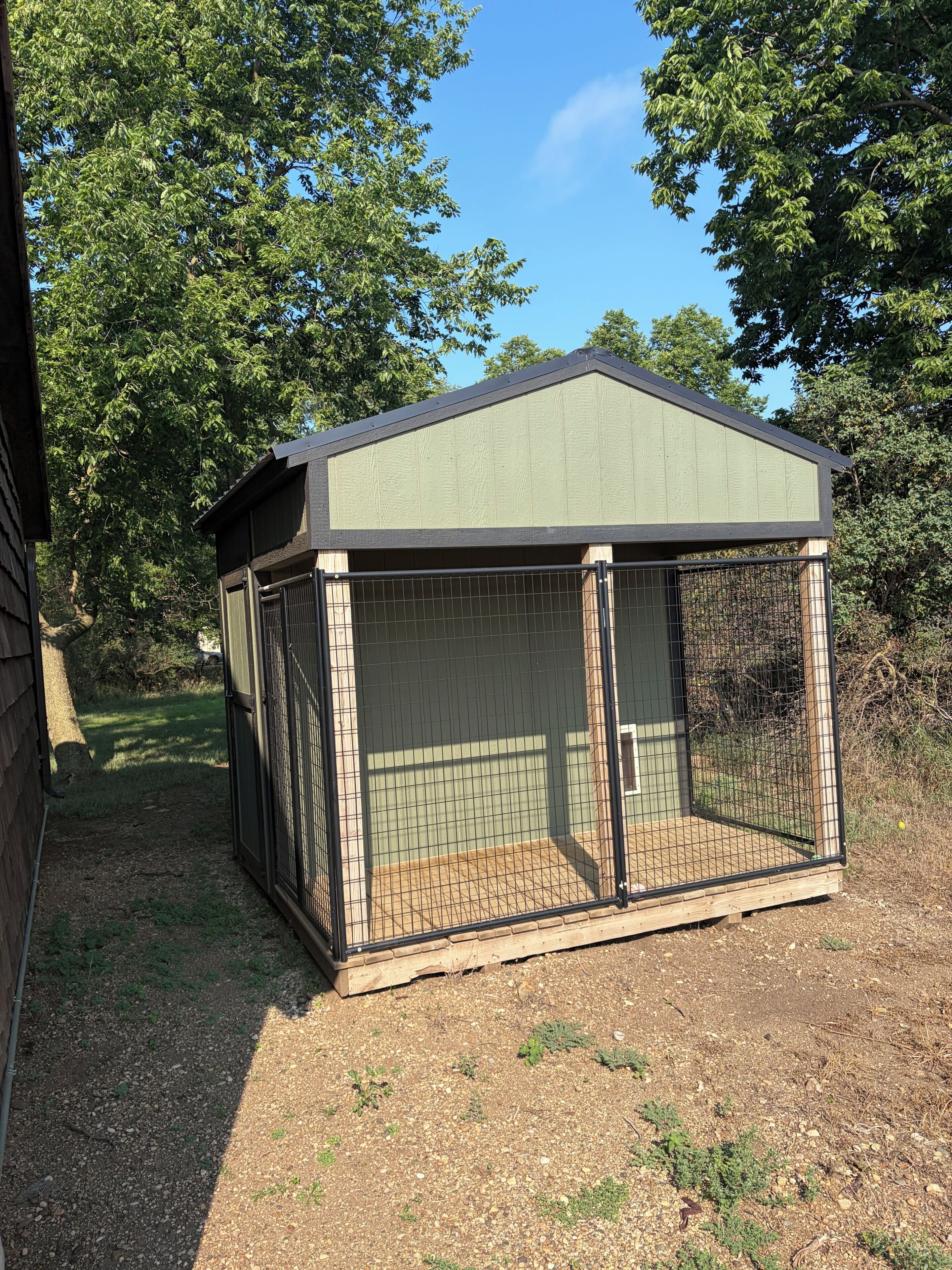 A fenced outdoor structure with a sloped roof, surrounded by trees.