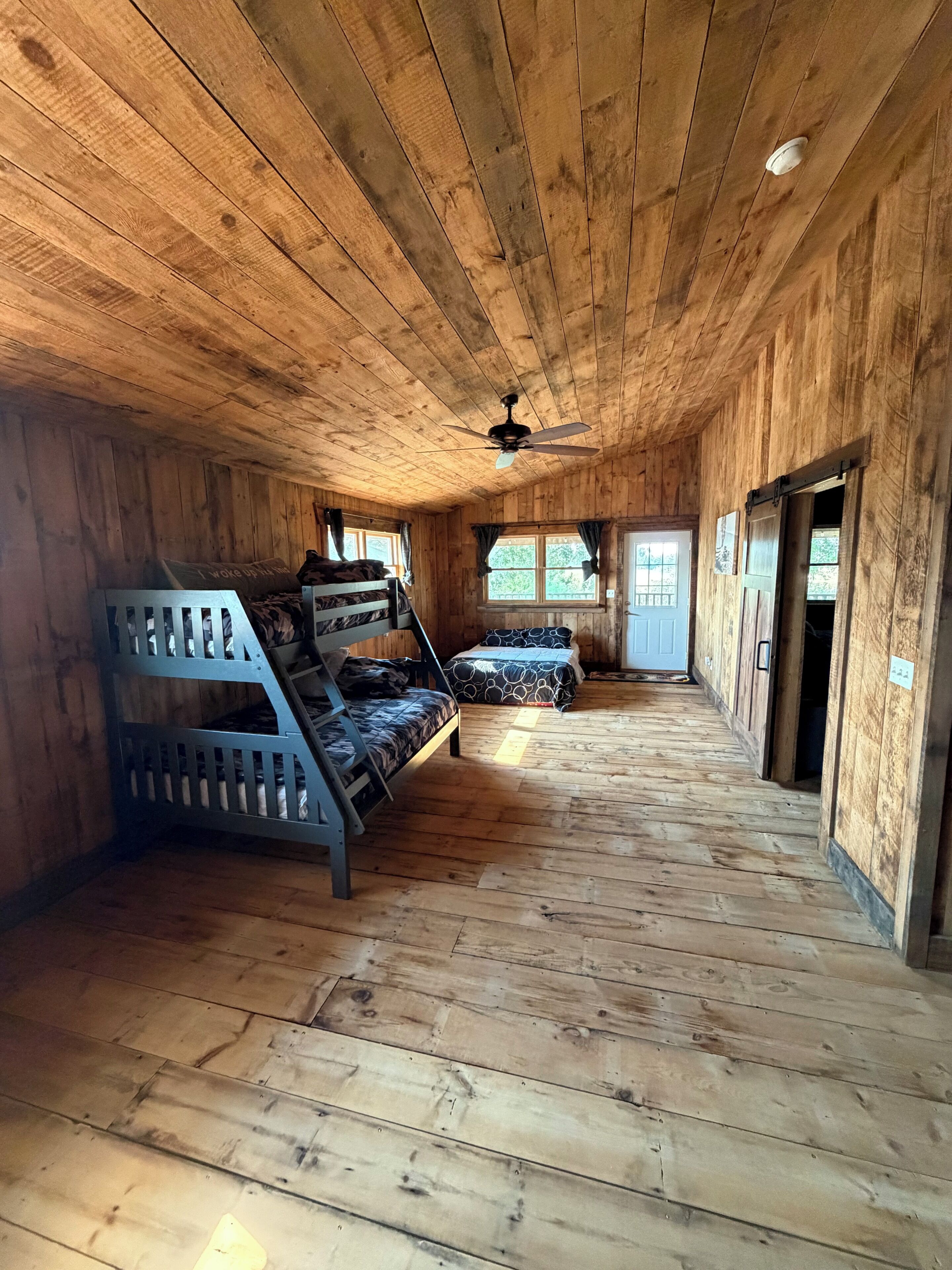 A wooden cabin interior with bunk beds, a ceiling fan, and a door leading outside.