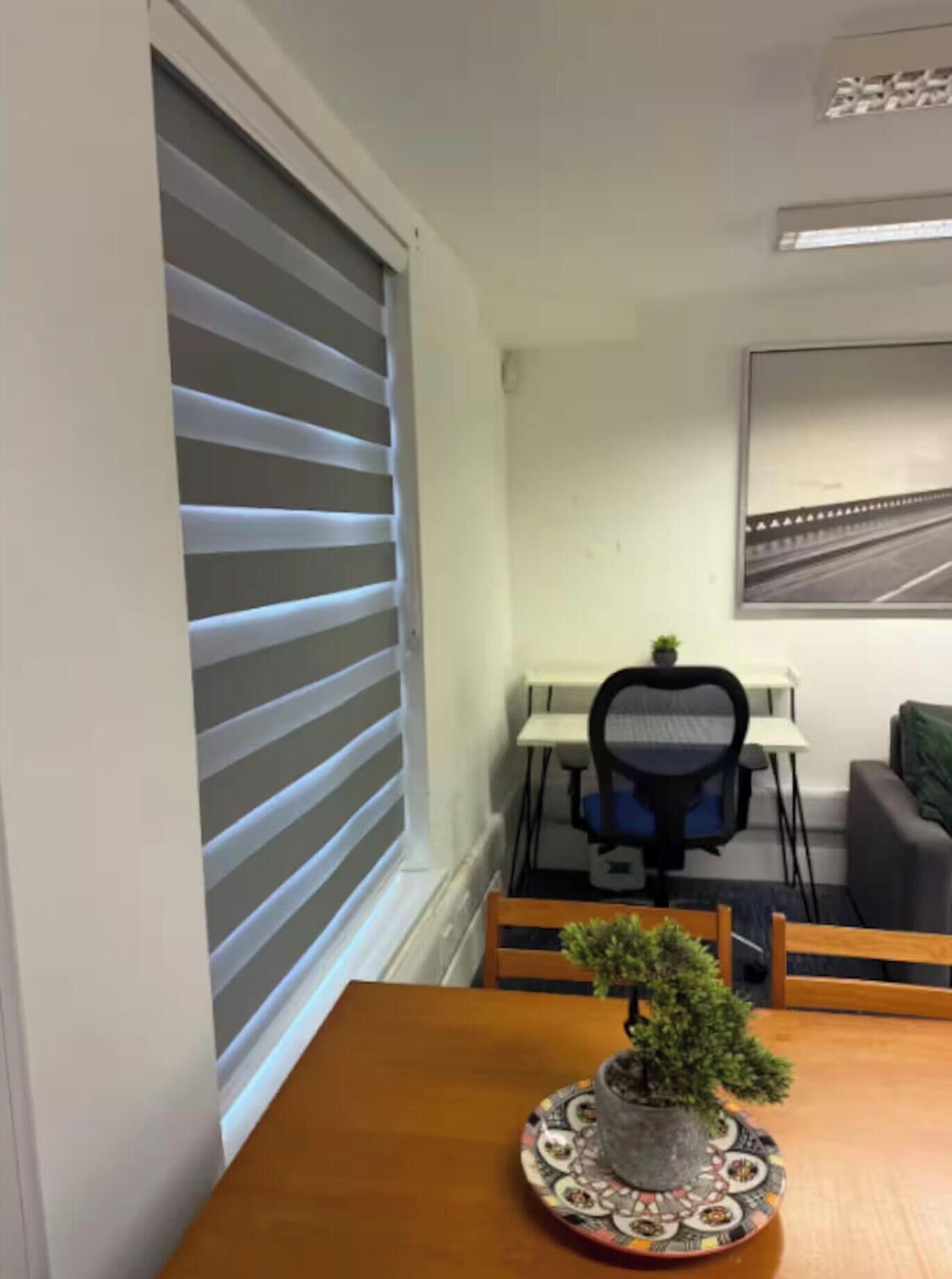 A room with a table, chairs, a potted plant, a desk with a chair, and a window with blinds.