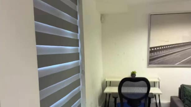 Ein Zimmer mit Tisch, Stühlen, einer Zimmerpflanze, einem Schreibtisch mit Stuhl und einem Fenster mit Jalousien.