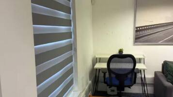 A room with a table, chairs, a potted plant, a desk with a chair, and a window with blinds.